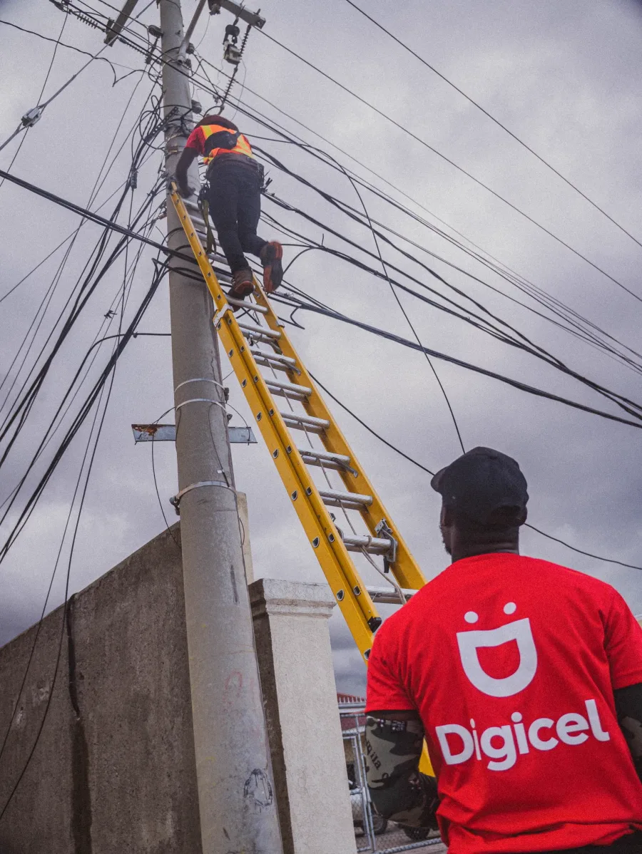 A Digicel technician on a ladder leaning against a pole, surrounded by many cables